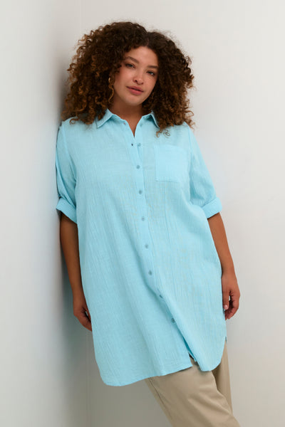 Billede af plus size model iført en lang lyseblå skjorte. Skjorten er gennemknappet og har en lomme, på det ene bryst. Ærmerne er smøget op, så de går til albuerne.  