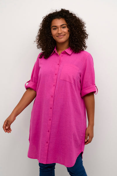 Plus size model med lang pink skjorte. Skjorten har en lomme på det ene bryst og er gennemknappet. Ærmerne er smøget op til albuerne. 